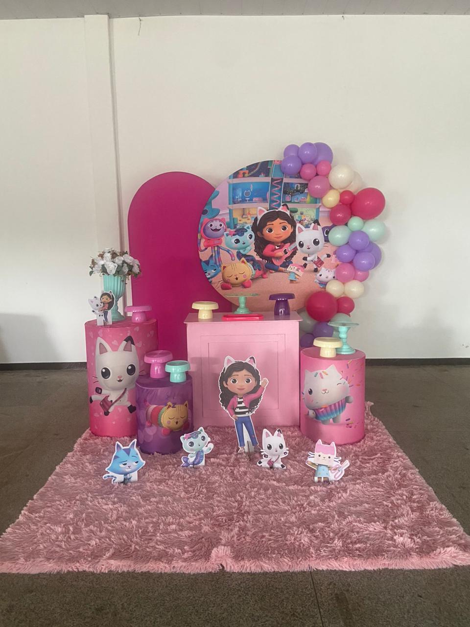Decoração tema Gabby's Dollhouse com painel redondo, cilindros rosa e lilás, tapete peludo e arco de balões candy colors
